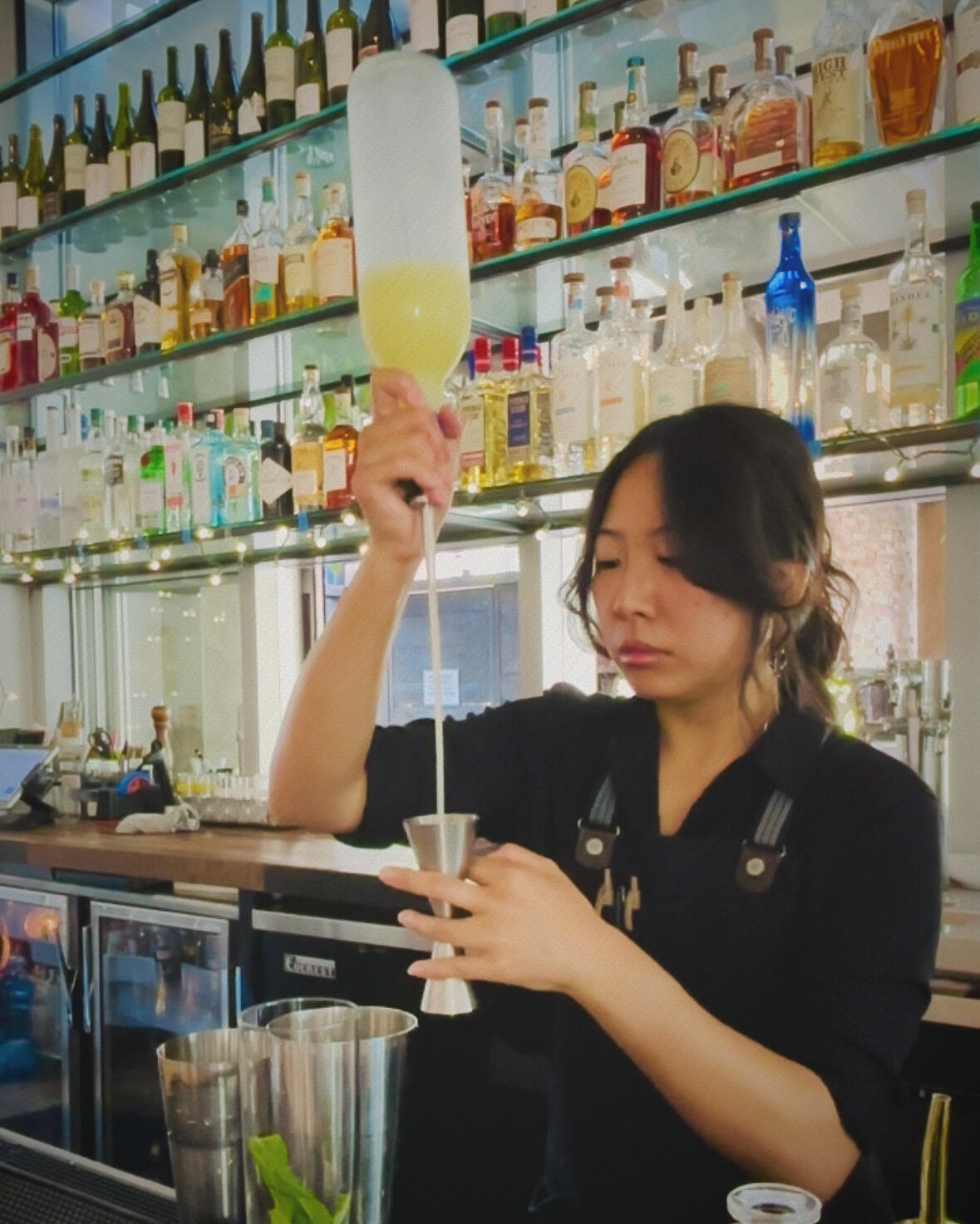 Alice - working at bar A woman with curtain bangs pouring lemon juice from a long-spout bottle into a jigger behind a bar.
